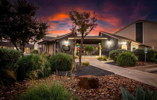 A building with a dark facade is surrounded by a garden with a large rock in the foreground.