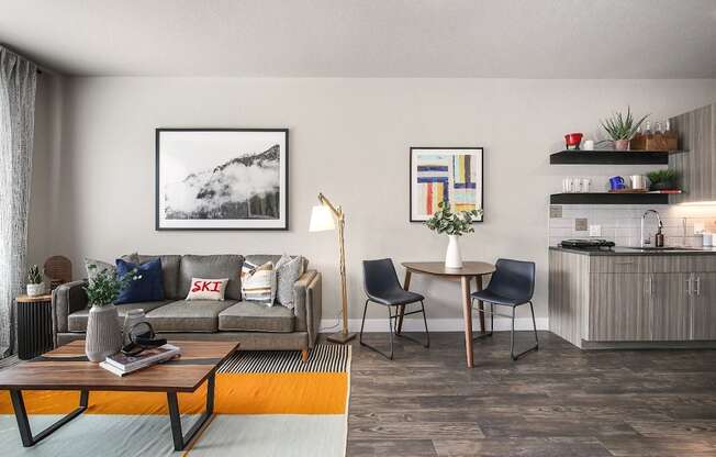 A modern living room with a grey sofa, a wooden coffee table, a grey armchair, a small dining table with two chairs, a bookshelf with various items, and a painting on the wall.