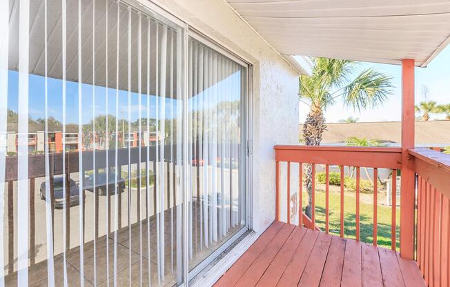 A balcony with wooden flooring and vertical blinds covering large glass sliding doors. The view outside includes palm trees and a parking area. The setting is bright and inviting, showcasing a well-maintained outdoor space.