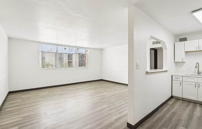 A room with white walls and wooden flooring, with a window and a small kitchenette.