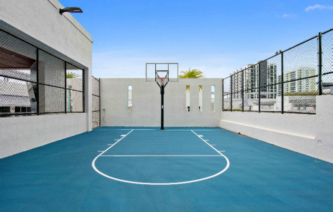Basketball court at our apartments in Miami, featuring blue asphalt, a plexiglass backboard, and black fences.