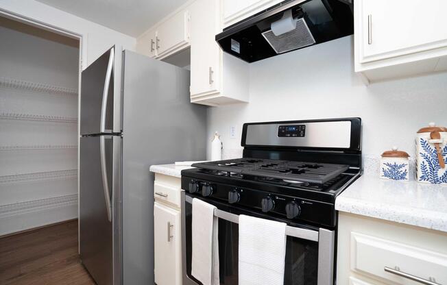 A modern kitchen featuring a stainless steel refrigerator, a gas stove with a black range hood, and white cabinets. The countertop is a light color, and there are two dish towels hanging in front of the stove. A pantry is visible in the background, with shelves ready for storage.