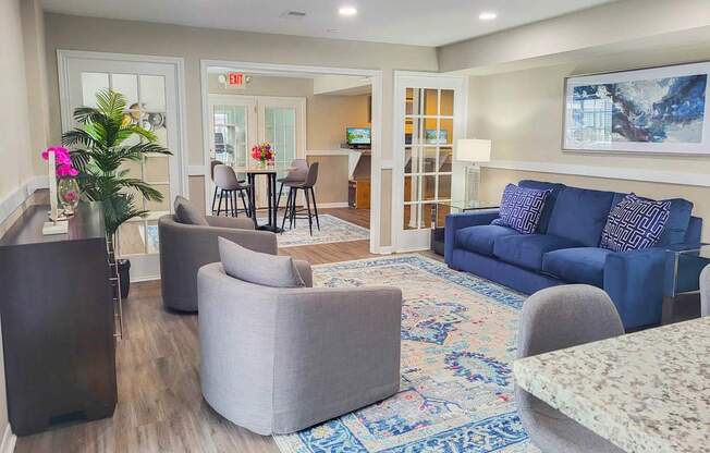 A living room with a grey sofa, a grey armchair, a grey ottoman, a wooden side table, a glass table with chairs, a rug, and a painting on the wall.