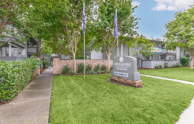 Sign for Creekside Apartments located on a manicured lawn, flanked by trees and shrubs. Two flags are visible, and the apartment buildings are in the background under a clear blue sky. A walkway leads towards the sign, enhancing the welcoming atmosphere of the complex.