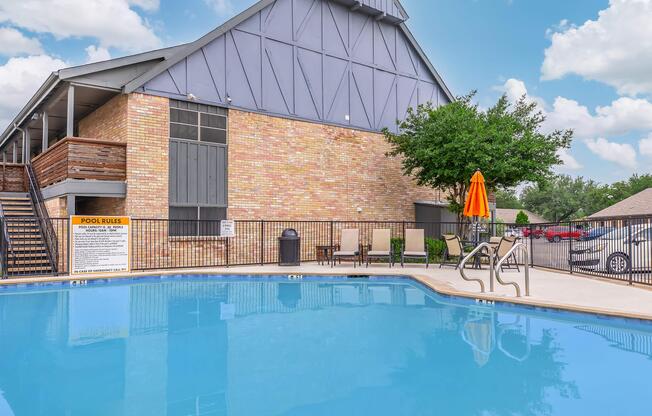 A clear blue swimming pool with a ladder in the foreground, surrounded by a black metal fence. A building with a brick facade and large windows is in the background, along with a green tree. An orange umbrella shades a seating area nearby, and pool rules are posted on a sign. The sky is partly cloudy.