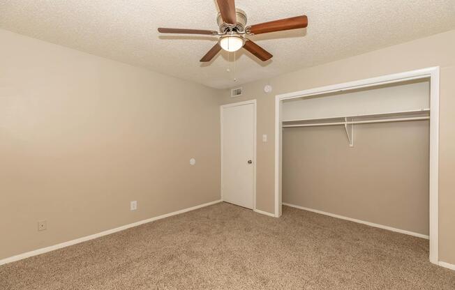 A spacious and neutral-colored bedroom featuring a ceiling fan, light-colored walls, and soft carpet. There is a closet with a single shelf and a door leading to another room. The room has a bright appearance, offering ample space for furniture placement.