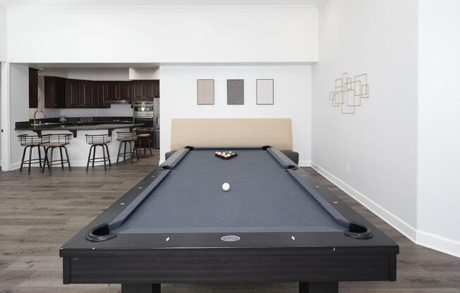 A pool table with a single white ball and a set of colored balls in a modern living space. Behind the table, there is a seating area and a kitchen with dark cabinetry. The walls feature simple artwork and decorative elements, creating a bright and contemporary atmosphere.