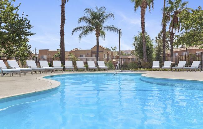 A clear blue swimming pool surrounded by lounge chairs and palm trees. In the background, residential buildings are visible against a bright blue sky. The scene conveys a relaxing outdoor atmosphere, ideal for leisure and sunbathing.