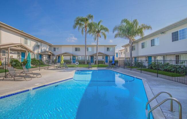 A bright and inviting swimming pool surrounded by lounge chairs and palm trees, with multiple apartment buildings in the background. The sky is clear and blue, enhancing the serene atmosphere of the outdoor space. Sunlight reflects off the water, creating a relaxing environment.
