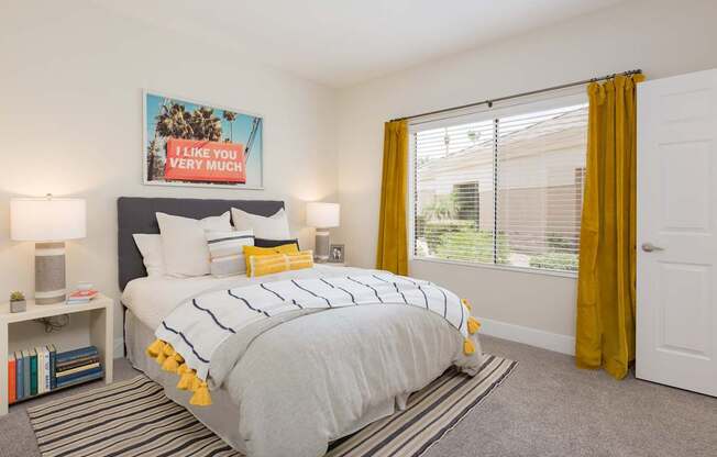 A bedroom with a bed, a nightstand, a lamp, a picture frame, a bookshelf, a rug, and a window with yellow curtains.