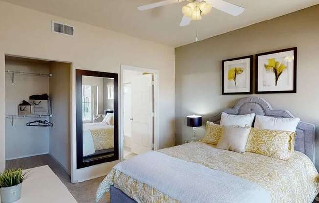 A spacious bedroom here at Dobson Towne Center featuring plush carpet, neutral walls, and a ceiling fan with light. A comfortable bed with decorative bedding anchors the room, while an open closet and full-length mirror add functionality. An adjoining bathroom enhances convenience and creates a private, well-designed retreat.