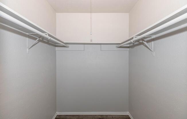 Empty closet with white walls and two horizontal hanging rods on either side. The floor is wooden, and there is a light fixture hanging from the ceiling. The space is well-lit and appears neatly organized, with no items or clothing present.