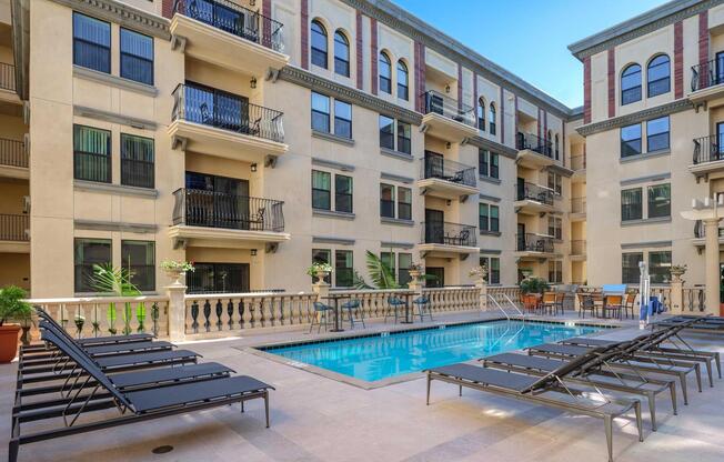 A serene courtyard featuring a clear swimming pool surrounded by lounge chairs. The area is enclosed by elegant apartment buildings with balconies, plants, and decorative details, creating an inviting atmosphere for relaxation and leisure. Bright sunlight enhances the overall ambiance.