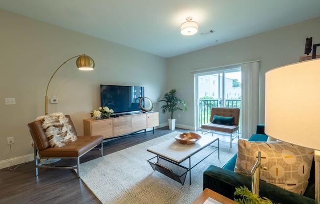 A bright living room with floor-to-ceiling sliding doors leading to a private balcony, furnished with modern seating and a flat-screen TV.