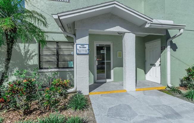 Entrance to a light green building with a covered entryway. The door has a sign indicating that it is open. There are plants and landscaping around the entrance, with a small patio area featuring decorative stone flooring. A window with blinds is visible to the left.