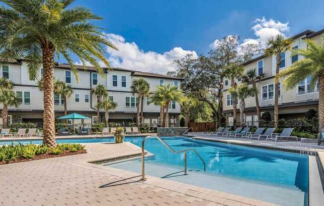 A swimming pool surrounded by palm trees and lounge chairs in front of apartment buildings.