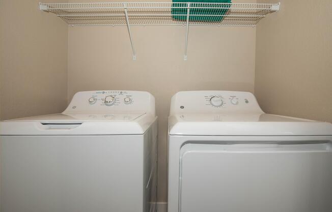 A laundry room featuring a top-loading washing machine and a front-loading dryer. Above them is a white wire shelf with a teal washcloth neatly placed on it, set against a simple beige wall. The floor is clean, and the overall appearance is tidy and organized.