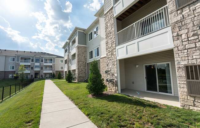 Paved sidewalk along the community, green grass, patios of apartment homes on right, fence next to pond on left.