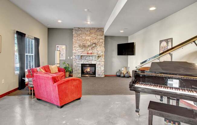 A stylish resident lounge here at Allegro featuring a striking floor-to-ceiling stone fireplace, plush red lounge chairs, and a polished concrete floor, complemented by a grand piano, wall-mounted television, and soft accent lighting that creates a warm, inviting space for relaxing, socializing, or hosting gatherings.