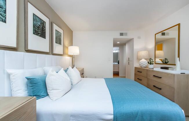 A modern bedroom featuring a large bed with white and turquoise bedding, decorative pillows, and a bedside lamp. The walls are adorned with framed botanical prints. A wooden dresser and a large mirror are present, along with soft lighting, creating a cozy and inviting atmosphere.