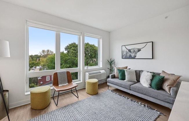 A living room with a grey couch, a yellow ottoman, a grey rug, a chair, a floor lamp, and a picture on the wall.