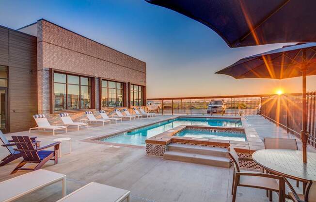 A sunset view of a pool area with lounge chairs and a building.