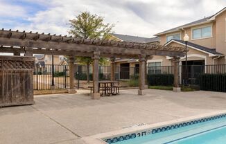 Swimming pool with tanning deck, pergola, picnic tables, and a green tree.