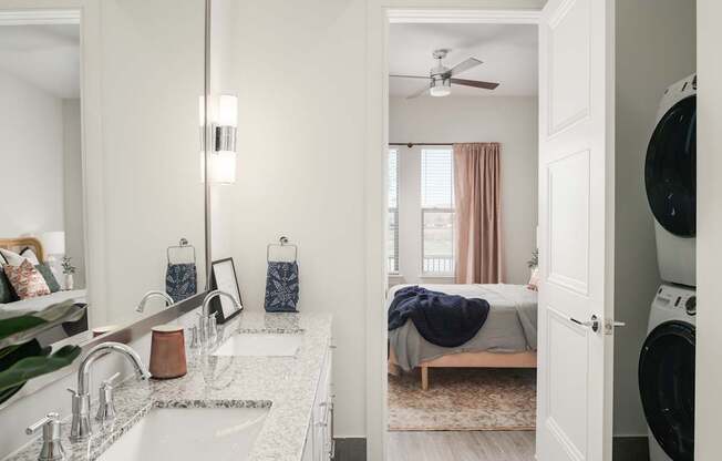 A modern bathroom with a double sink vanity and a view into a bedroom.