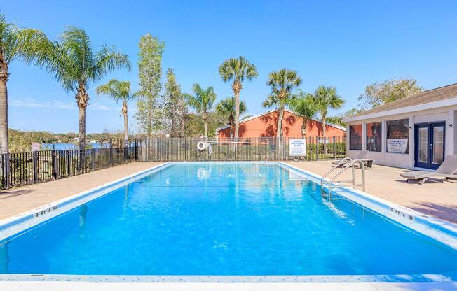 A clear blue swimming pool surrounded by palm trees, with a fenced area and lounge chairs. In the background, there is a vibrant orange building and a bright blue sky, creating a relaxing outdoor atmosphere.