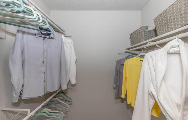 A neatly organized closet featuring a variety of clothing on hangers, including a white shirt, a light blue shirt, a yellow sweater, and a gray top. There are also woven storage baskets on a shelf above, and empty hangers are visible, indicating available space for more items. The walls are plain and white.