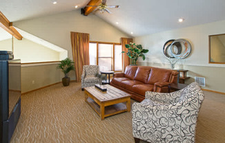 A spacious living room here at Eagle Ridge showcasing vaulted ceilings with exposed wood beams, a leather sofa centered around a wooden coffee table, patterned accent chairs, soft carpeted flooring, and large windows with warm curtains that bring in natural light and create an inviting, comfortable gathering space.