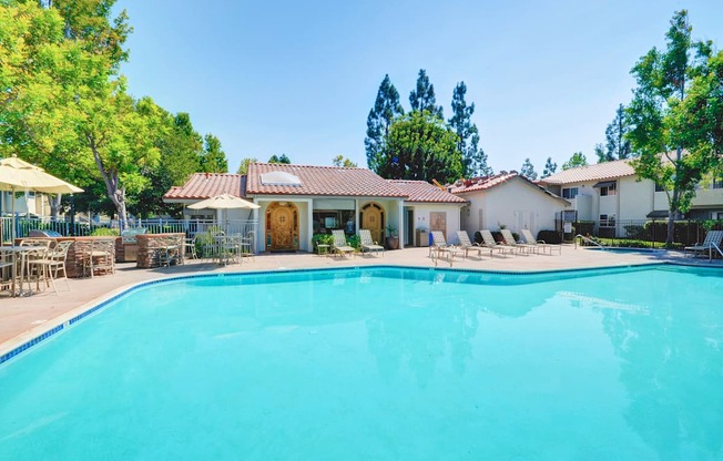A large swimming pool in front of a house with a patio and trees in the background.