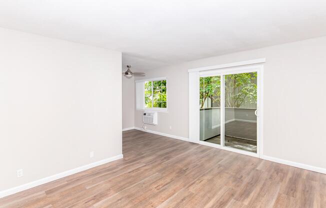 Spacious and well-lit living room featuring light wood laminate flooring, neutral-colored walls, and large sliding glass doors leading to a balcony. A window provides natural light, and a ceiling fan is installed for ventilation. An air conditioning unit is visible on the wall.