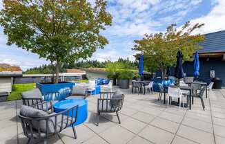 A patio with blue chairs and tables is surrounded by trees. at Kirkland Crossing Apartments, Kirkland, WA