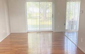 A bright living room with a sliding glass door that leads to the outdoors. The floors are shining wood and the walls are light colored.