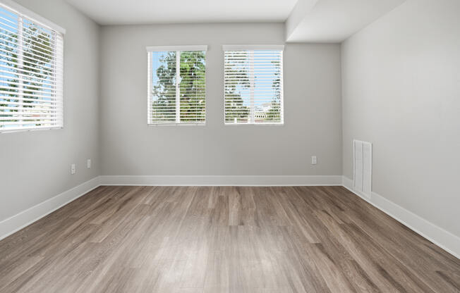 A room with wooden flooring and two windows with blinds.