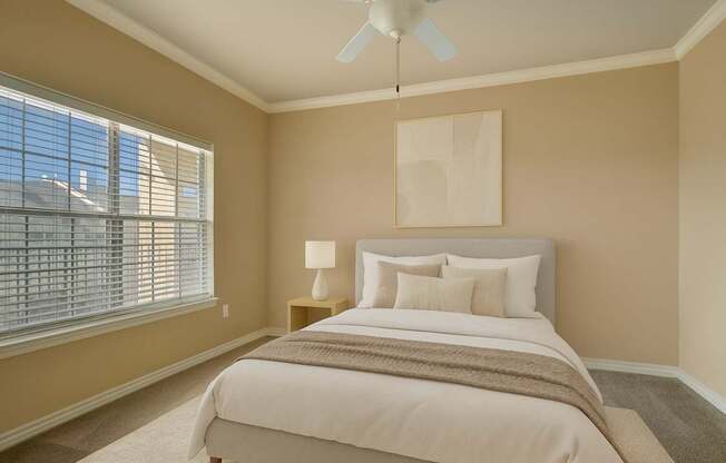 A cozy and serene bedroom here at Mission Green featuring a neatly made bed with white bedding, layered neutral pillows, and a soft throw blanket, complemented by warm beige walls, plush carpeting, a ceiling fan with light, simple abstract wall art above the headboard, and natural light filtering through wide window blinds.