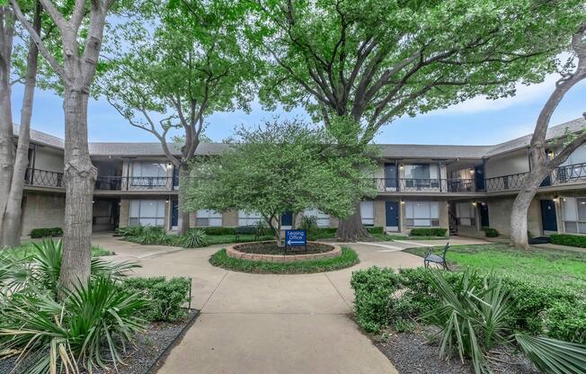 A courtyard view of an apartment complex featuring lush green trees, well-maintained landscaping with shrubs and grass, and two-story buildings with balconies. A sign indicating "Garden Apartments" is visible. The scene is peaceful with a clear sky, creating a welcoming atmosphere.