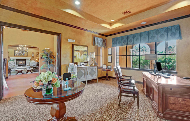 A refined business center here at Tuscany at Faudree with a large wooden desk, guest chairs, and a computer workstation. Decorative tables, framed artwork, and soft carpeting add an upscale feel, while large windows bring in natural light and connect the space to adjacent community lounges.