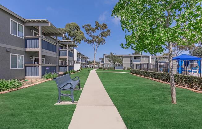 A neat pathway lined with green grass leads between modern apartment buildings. Benches are placed along the path, and trees provide shade. In the background, there's a blue pool area, surrounded by fencing and more greenery, creating a welcoming community atmosphere.