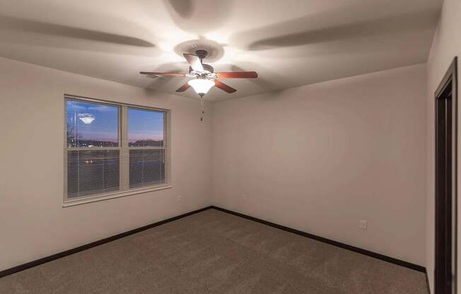A cozy bedroom here at The Park featuring soft carpet, a bright window with blinds, neutral walls, and a ceiling fan, creating a calm and inviting space.