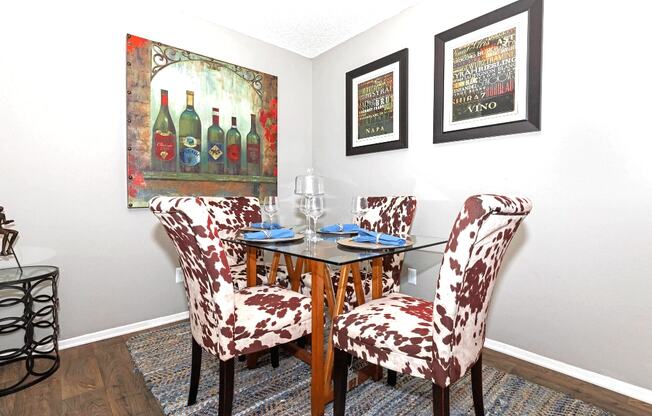 A dining room with a glass table and chairs with a wine bottle painting on the wall.