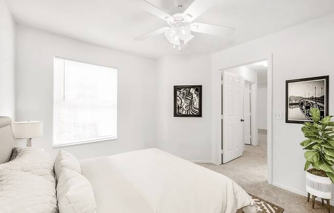 Virtually staged bedroom with carpet, white walls, ceiling fan with light, large window, accent rug, and plant decor.