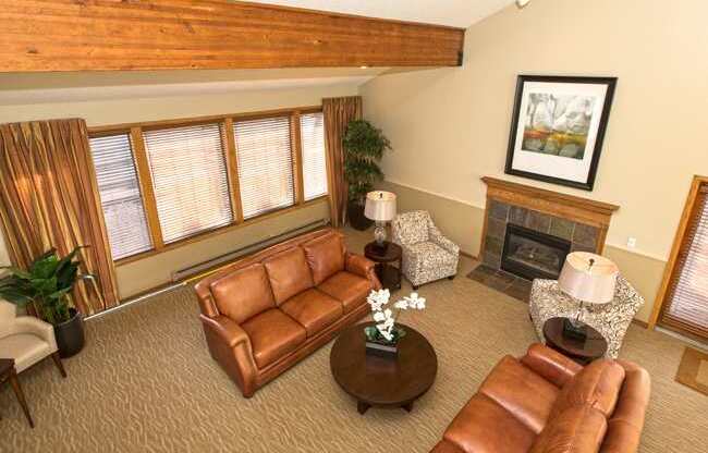 A bright, spacious living room here at Eagle Ridge featuring vaulted ceilings with exposed wood beams, warm neutral walls, leather sofas arranged around a round coffee table, patterned accent chairs, a cozy fireplace with a wood mantel, soft carpeted flooring, and large windows with blinds that allow natural light to fill the inviting space.