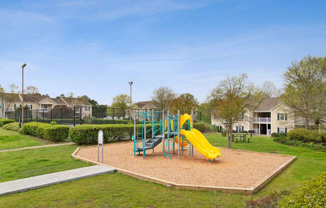 Playground in bed of mulch and yellow slide with a sports court in the back