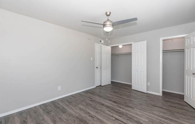 A clean, empty room with light gray walls and a ceiling fan. The floor is made of light wood laminate. There are two open white doors leading to closets on the right, and a single door on the left. The room has no furniture or decorations, creating a spacious and bright atmosphere.