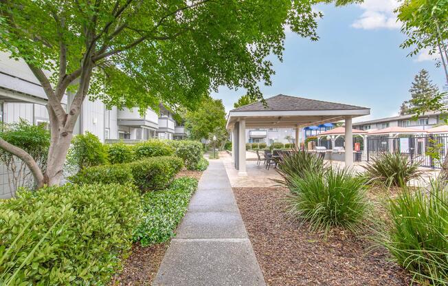 View of a tree-lined pathway leading to a gazebo surrounded by lush greenery. The path is lined with shrubs and ornamental grasses, and there are residential buildings visible in the background. A swimming pool area is partially visible, adding to the serene outdoor ambiance.