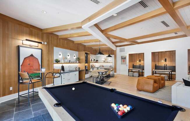 A stylish community lounge here at Reflect at Dobson Ranch showcasing a modern game and gathering space with a billiards table in the foreground, warm wood-paneled walls, coffered ceilings with exposed beams, a fully equipped kitchenette, high-top seating, built-in work alcoves, and comfortable lounge seating designed for socializing and relaxation.