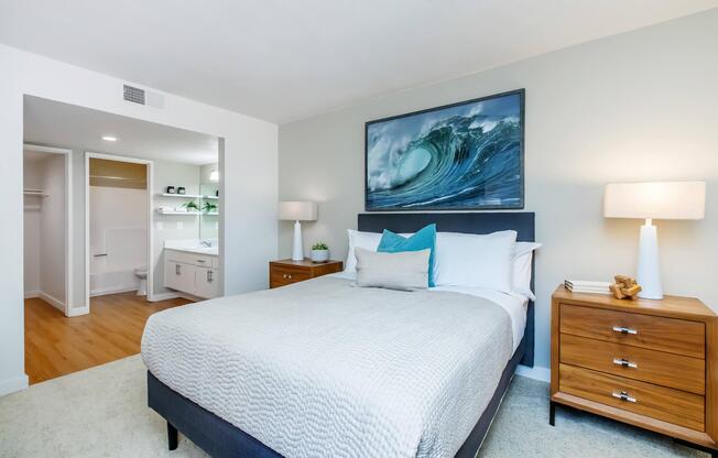 A cozy bedroom featuring a queen-sized bed with light bedding, two wooden nightstands on either side, a large wave-themed artwork above the bed, and soft lighting from lamps. An adjoining bathroom with modern fixtures is visible in the background, showcasing a clean and inviting atmosphere.