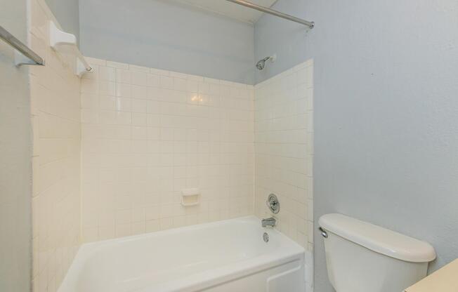 A clean, simple bathroom featuring a white bathtub with a showerhead, white tile walls, and a toilet nearby. The walls are painted light blue, and there's a towel bar mounted on the wall. Natural light from a nearby window illuminates the space.